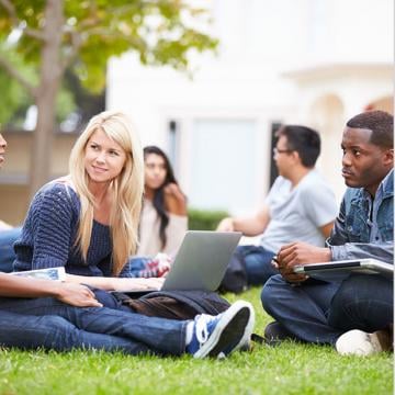Un grupo diverso de estudiantes sentados en el césped, absortos en sus computadoras portátiles, estudiando y colaborando al aire libre.