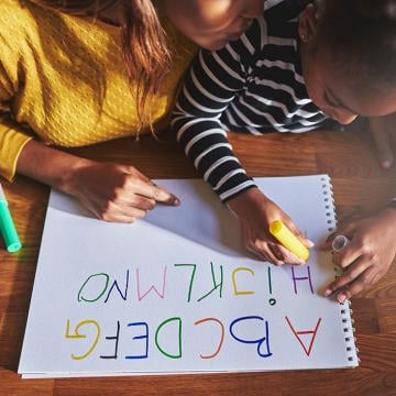 Una mujer y un niño sentados en una mesa y el niño está aprendiendo a escribir alfabetos usando varios colores.