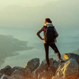 Una mujer con una mochila se para triunfalmente en la cima de una majestuosa montaña, abrazando la impresionante vista.
