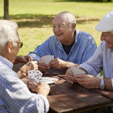 Tres ancianos disfrutando de una partida de cartas en el parque, rodeados de naturaleza y risas.